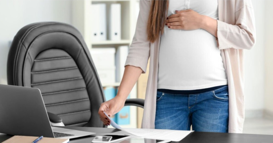 A pregnant woman stands at her desk, hands on her belly, reviewing papers.